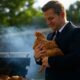 A man in a suit holding a live chicken while standing next to a smoky grill with chicken pieces cooking, set against a backyard background.
