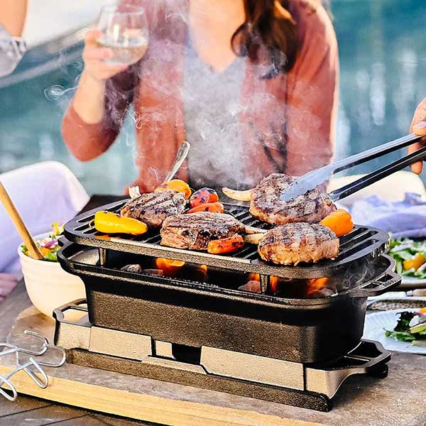 A close-up of a Lodge Sportsman Grill with sizzling food on the cast iron grate, with a person holding tongs and another enjoying wine in the background.