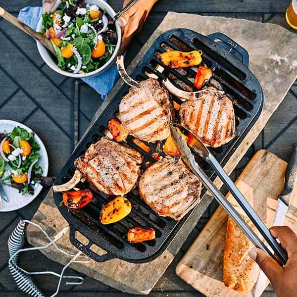 An overhead close-up of a Lodge Sportsman Grill with sizzling food on the cast iron grate, with a person holding tongs.