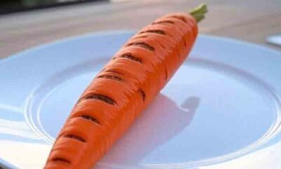 A single large grilled carrot with grill marks on a white plate, set on a wooden table near the ocean at sunset, featured on PopularBBQ.com