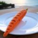 A single large grilled carrot with grill marks on a white plate, set on a wooden table near the ocean at sunset, featured on PopularBBQ.com