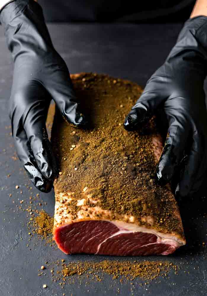 Hands applying brisket dry rub technique: Pressing coarse spices evenly into trimmed meat for adhesion and flavor penetration before smoking
