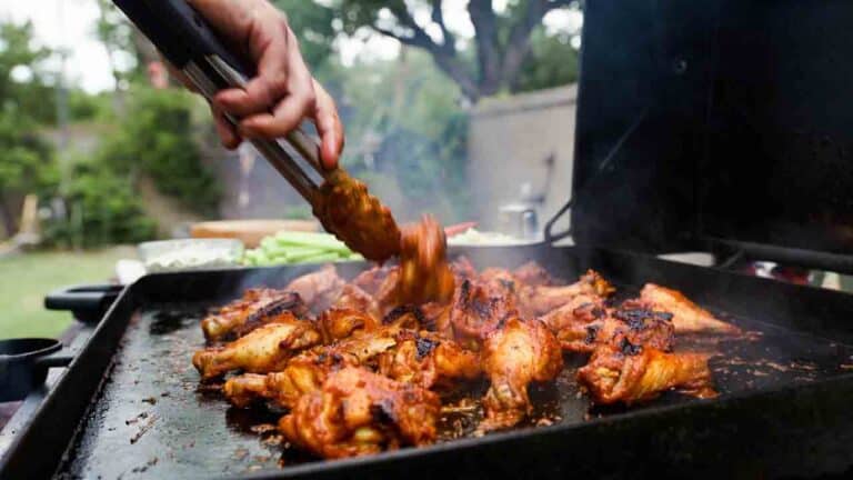 Golden crispy grilled buffalo chicken wings being tossed in tangy red sauce on an outdoor griddle, with celery sticks and blue cheese dip in a sunny backyard setting