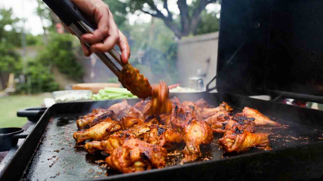 Grilled Buffalo Chicken Wings on the Griddle Hero Image Golden crispy grilled buffalo chicken wings being tossed in tangy red sauce on an outdoor griddle, with celery sticks and blue cheese dip in a sunny backyard setting
