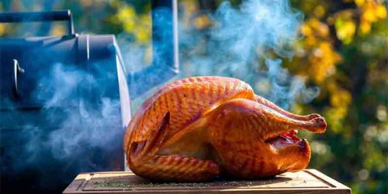 Whole smoked thanksgiving turkey with golden-brown crispy skin and pink smoke ring on a wooden board beside a smoker, blue smoke wisps, fall herbs and foliage background