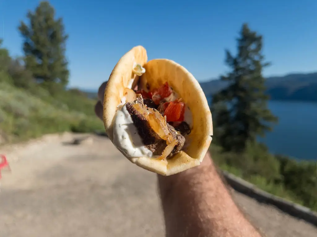 Bear_Souvlaki_Gyros_Simple_Gourmet_Levi_Sim-4-1024x768jpg - Popular BBQ Close-up of a pita sandwich with grilled meat and vegetables, held outdoors with a lake and trees in the background.