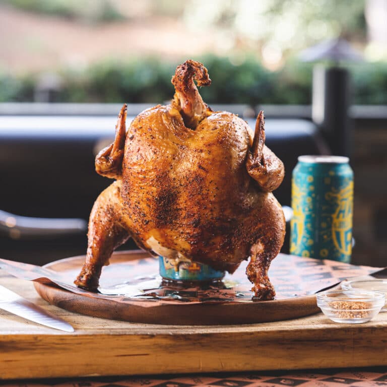 Whole roasted chicken perched on a beer can, crispy skin, on a wooden board outdoors.