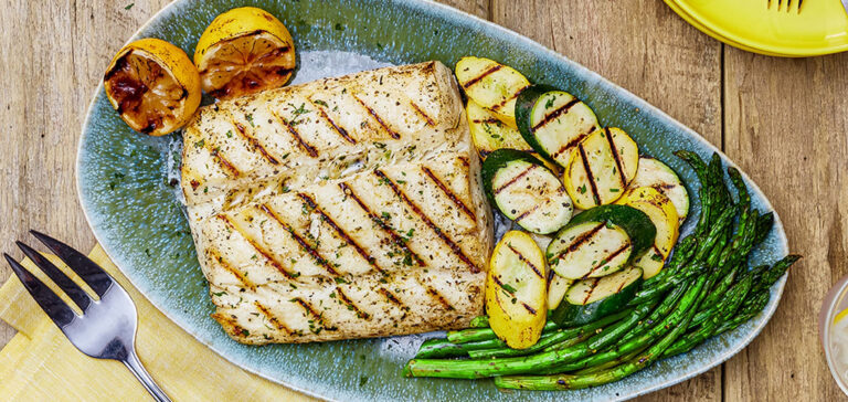 Grilled white fish fillet with lemon, potato slices, zucchini, and asparagus on a blue oval plate.