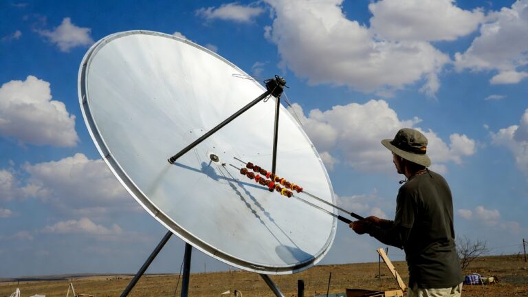 Man wearing a wide-brim hat adjusts a large satellite dish outdoors in a rural field under a blue sky with clouds.