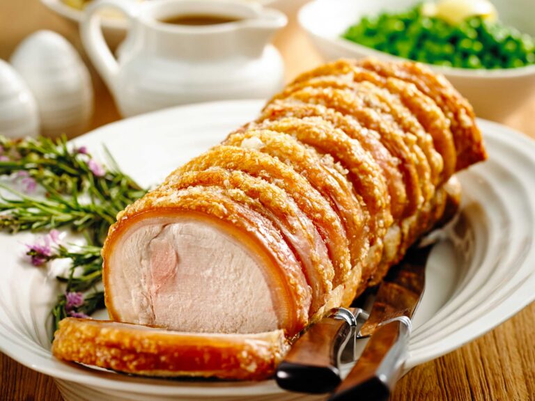 Sliced roasted pork with crispy crackling on a white plate, garnished with herbs and a carving knife nearby