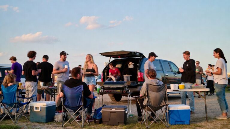 A group of people tailgating beside an SUV with a grill, coolers, and folding chairs under a clear sky.