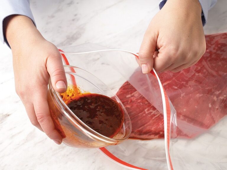 Hands pouring red marinade into a vacuum-seal bag containing raw steak on a marble surface.