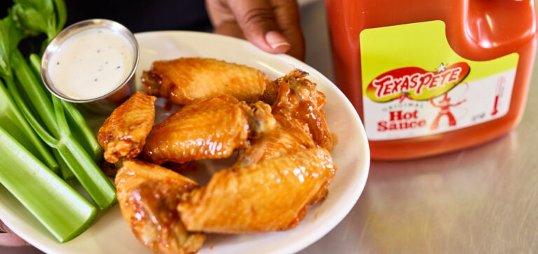Plate of spicy chicken wings with celery sticks and ranch dip, with a Texas Pete hot sauce bottle nearby.