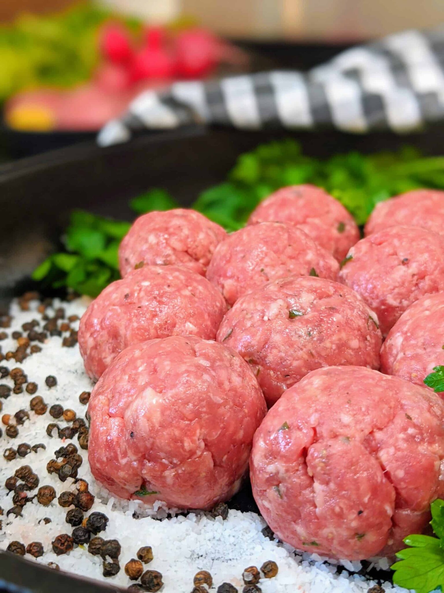 00100trPORTRAIT_00100_BURST20200911141126015_COVER-scaled - Popular BBQ Pile of pink raw meatballs seasoned with herbs on a bed of coarse salt, with scattered peppercorns and green garnish nearby
