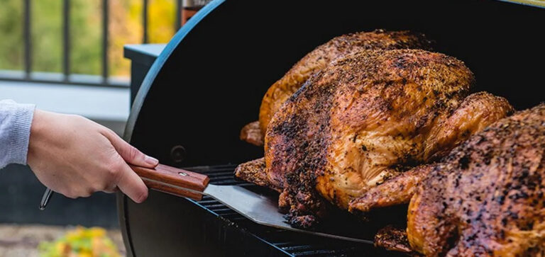Person carving a large smoked turkey on a grill with a wooden-handled knife in the foreground.